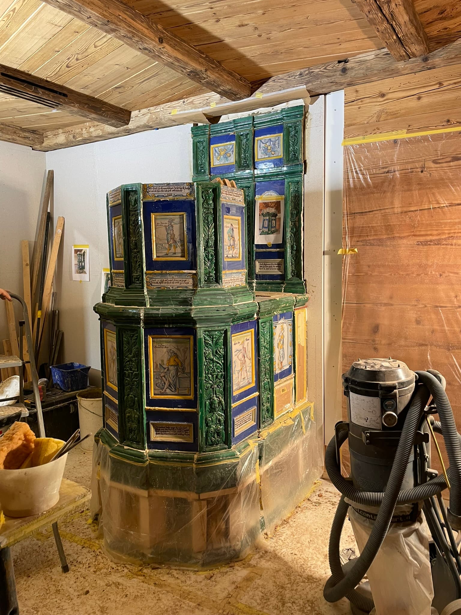 Wiederaufgebauter grün-blauer Ofen in einem Raum mit Holzbalken